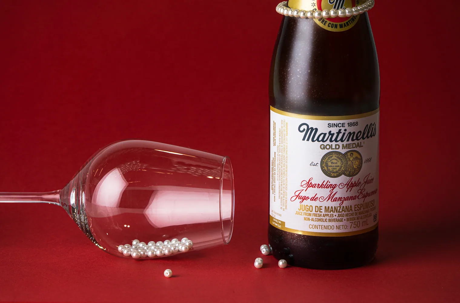 Botella de sidra espumosa y copa caída con perlas sobre fondo rojo
